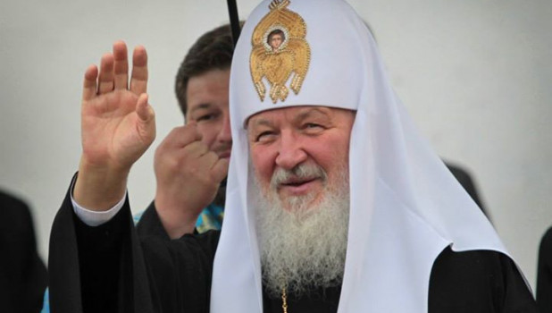 Охранник ФСО держит зонт над патриархом Кириллом.