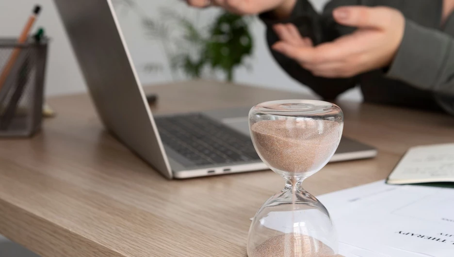 Женщина работает за ноутбуком, рядом стоят песочные часы.